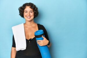 Young, smiling woman holding a yoga mat and towel, ready to exercise. Physicians recommend yoga and other exercises after surgery, especially for recovery following knee replacement.