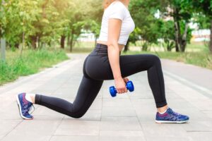 Woman performing a forward lunge outdoors while holding a dumbbell, demonstrating strength training for the lower body.