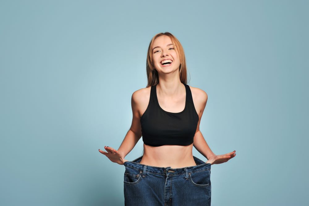 Happy woman wearing a loose trouser, holding the waist to show how much weight she’s lost after following a weight loss training program, smiling in an exercise top.