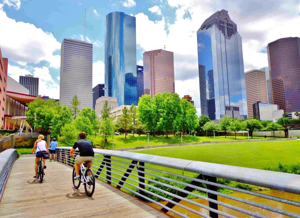 Bicyclists crossing a wooden bridge in Buffalo Bayou Park with a scenic downtown Houston skyline during Houston Holidays.