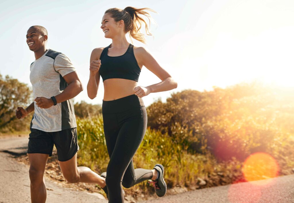 Young smiling couple jogging on a nature trail, illustrating the connection between regular exercise, happiness, and long-term health.