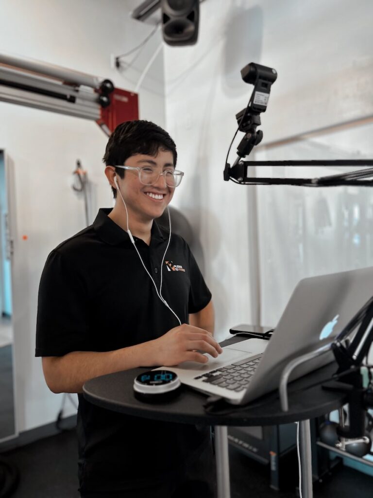 Online personal trainer at Pledge To Fitness in Bellaire conducting a virtual session using a laptop and headset in a professional studio setting.
