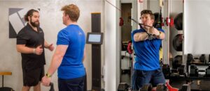 A personal trainer at Pledge To Fitness®'s Bellaire studio discusses body scan results with a client using the Fit3D scanner; the same client performs a resistance cable exercise as part of his personalized workout in the studio's private training pod.