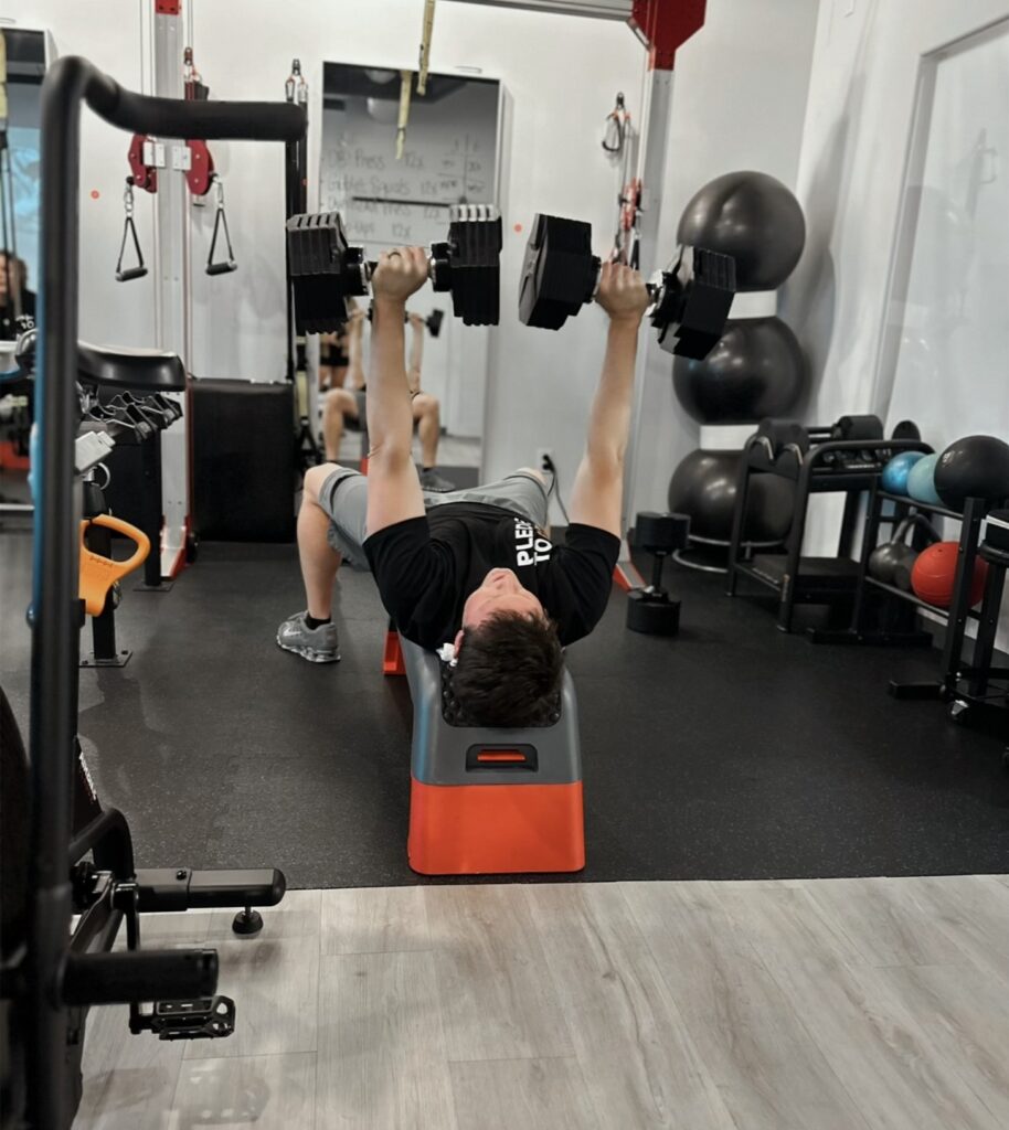 Man performing dumbbell bench press on a step bench at Pledge To Fitness® studio, focusing on strength training after 50.