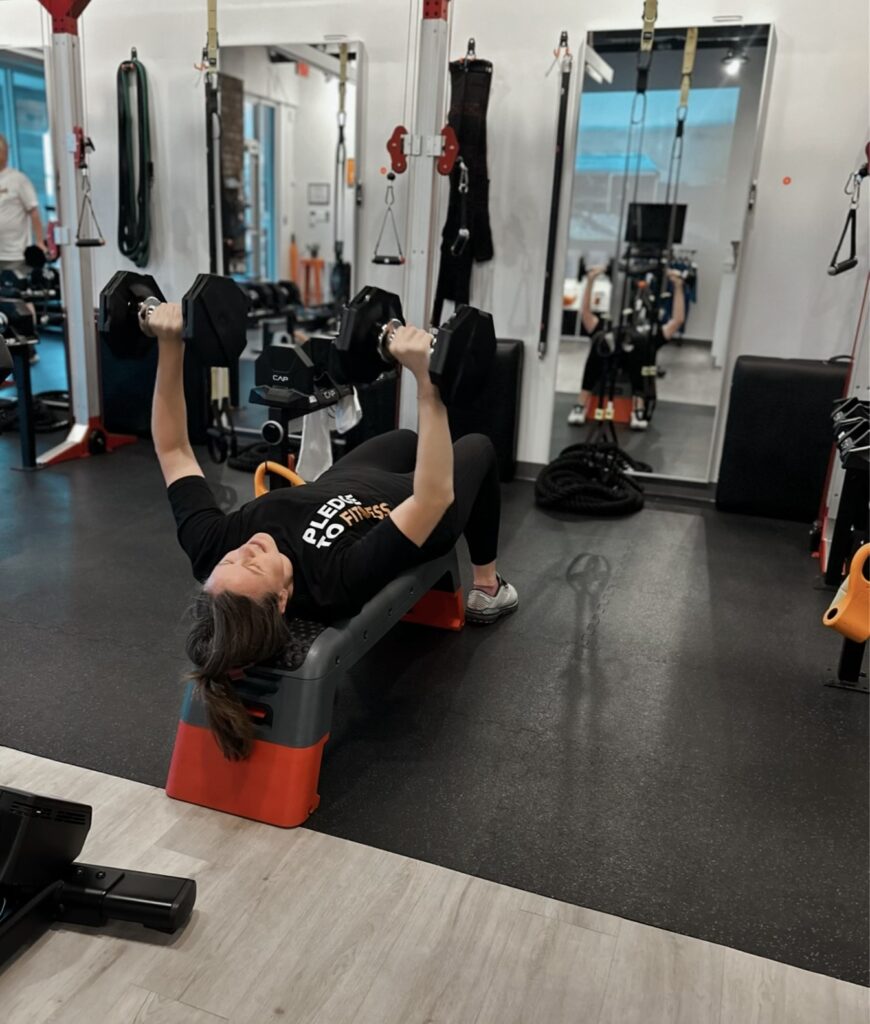 Woman performing a dumbbell bench press on a step bench at Pledge To Fitness® studio, demonstrating strength training after 50.