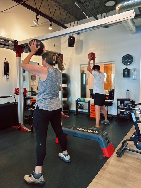 Couple training together at Pledge To Fitness® studio, performing overhead medicine ball exercise to build strength and lose weight after 50.